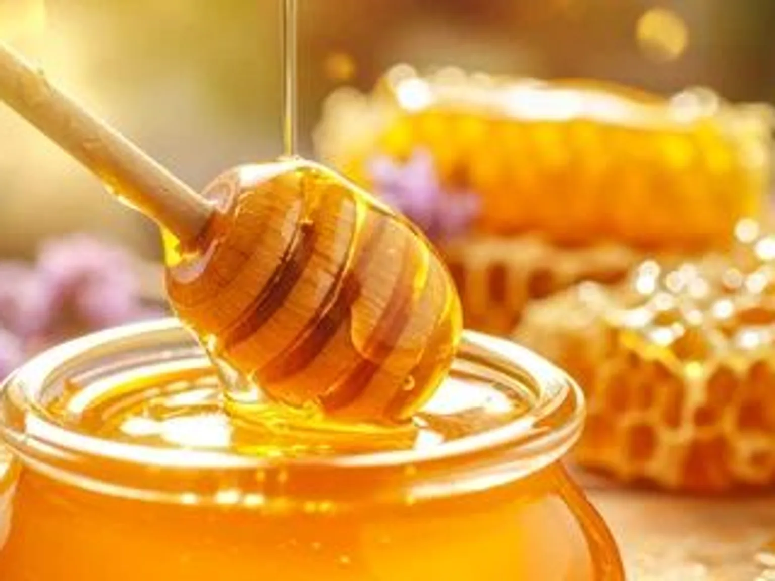 A wooden dipper lifting honey over an open glass jar, with honeycomb behind it.